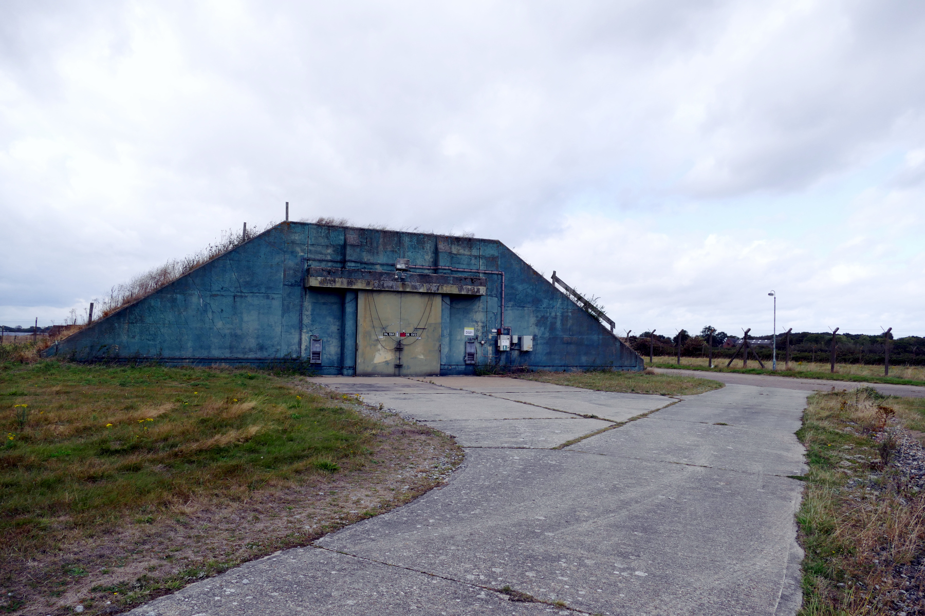 Wide shot of original Cold War bunker with blue walls, green blast door on grass with concrete road